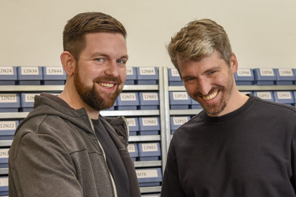 Zwei Kollegen lächeln in Werkstattbereich mit Regalen im Hintergrund