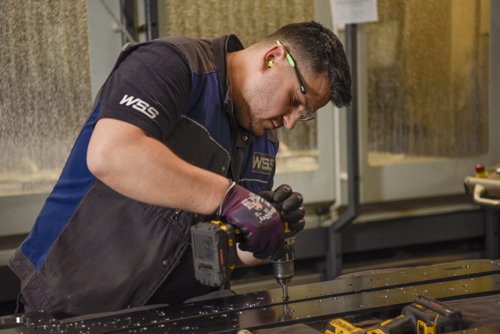 Mitarbeiter mit Schutzbrille bohrt durch eine lange glänzende Metallplatte